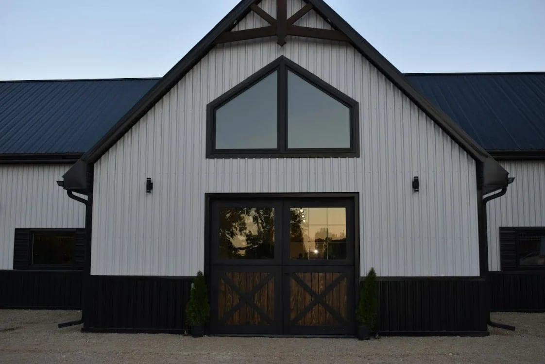 Modern farmhouse with white corrugated metal siding, black roof, large gable window, and barn-style wooden doors at dusk.